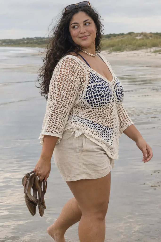 Curvy woman in a white crochet tunic and linen shorts on the beach, a plus size boho summer look